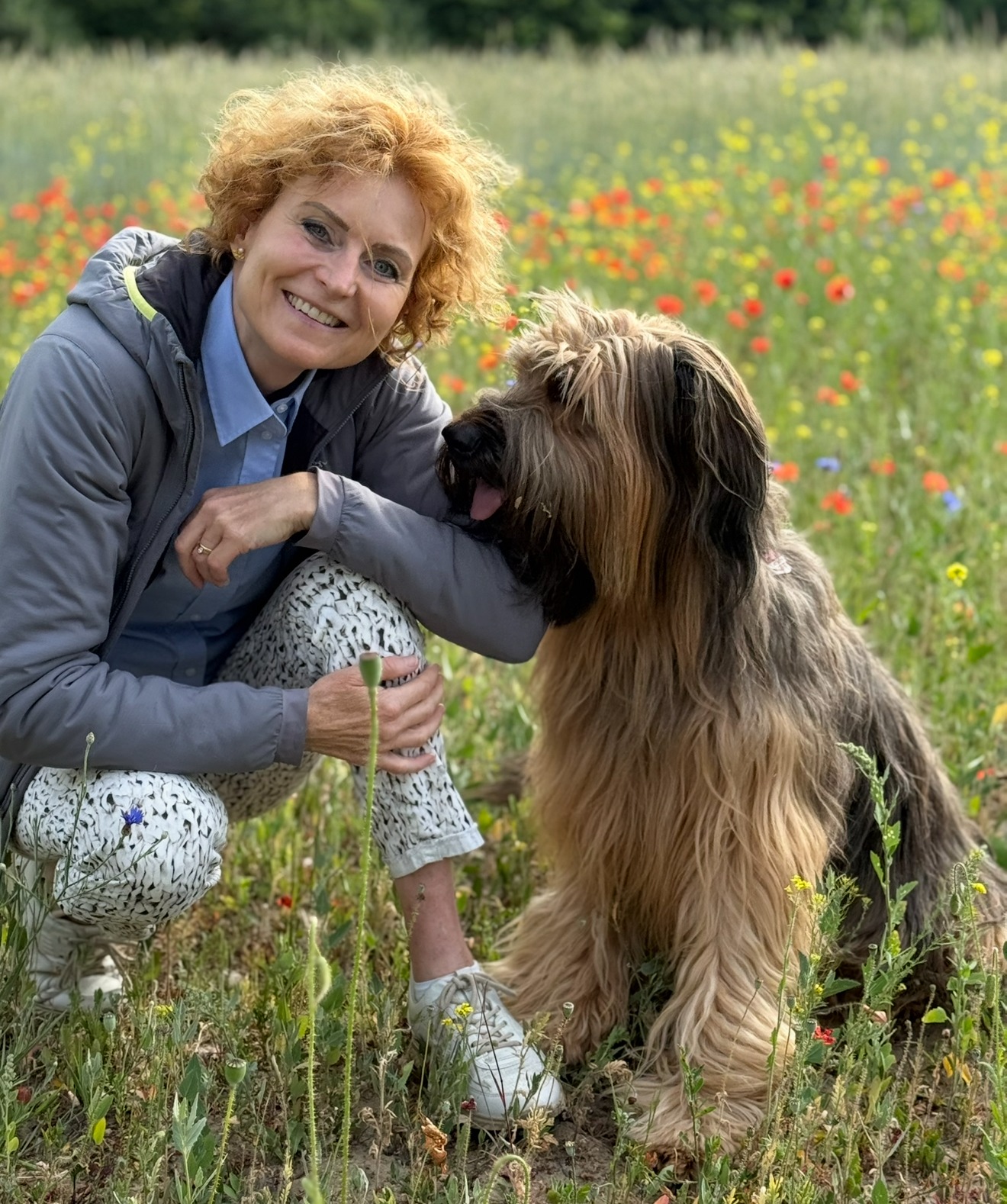 Frau mit Hund sitzen auf einer wild blühenden Wiese. Beide sind sich sehr nah. Die Frau lacht in die Kamera