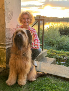 Coachin Antje Timmermann hockt mit ihrem Hund auf einer Mauer. Im Hintergrund sieht man Wasser mit Abenddämmerung - Philosophie Mind & Lead